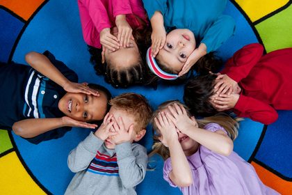 Niños de preescolar recostados en círculo sobre un tapete de colores, tapándose los ojos y riendo