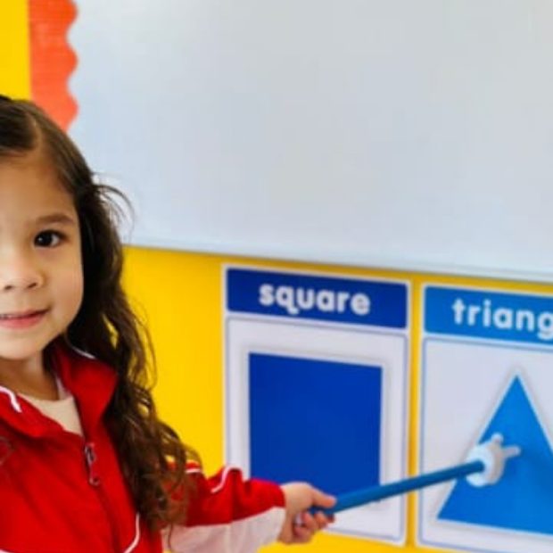 Niña de preescolar señalando con un puntero las tarjetas de figuras geométricas en inglés: square, triangle, rectangle
